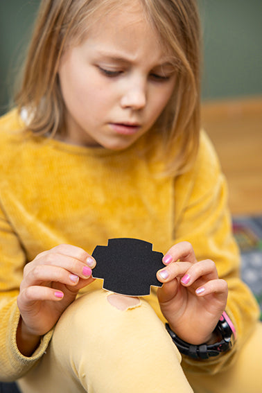 A child in a yellow outfit examines a FabPatch rub-on textile repair patch while looking at a tear in their yellow pants