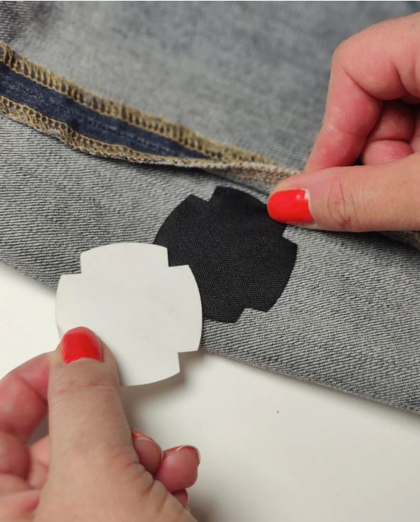 Hands with red nail polish applying a black FabPatch to repair a tear on denim fabric, showcasing a quick and easy fix