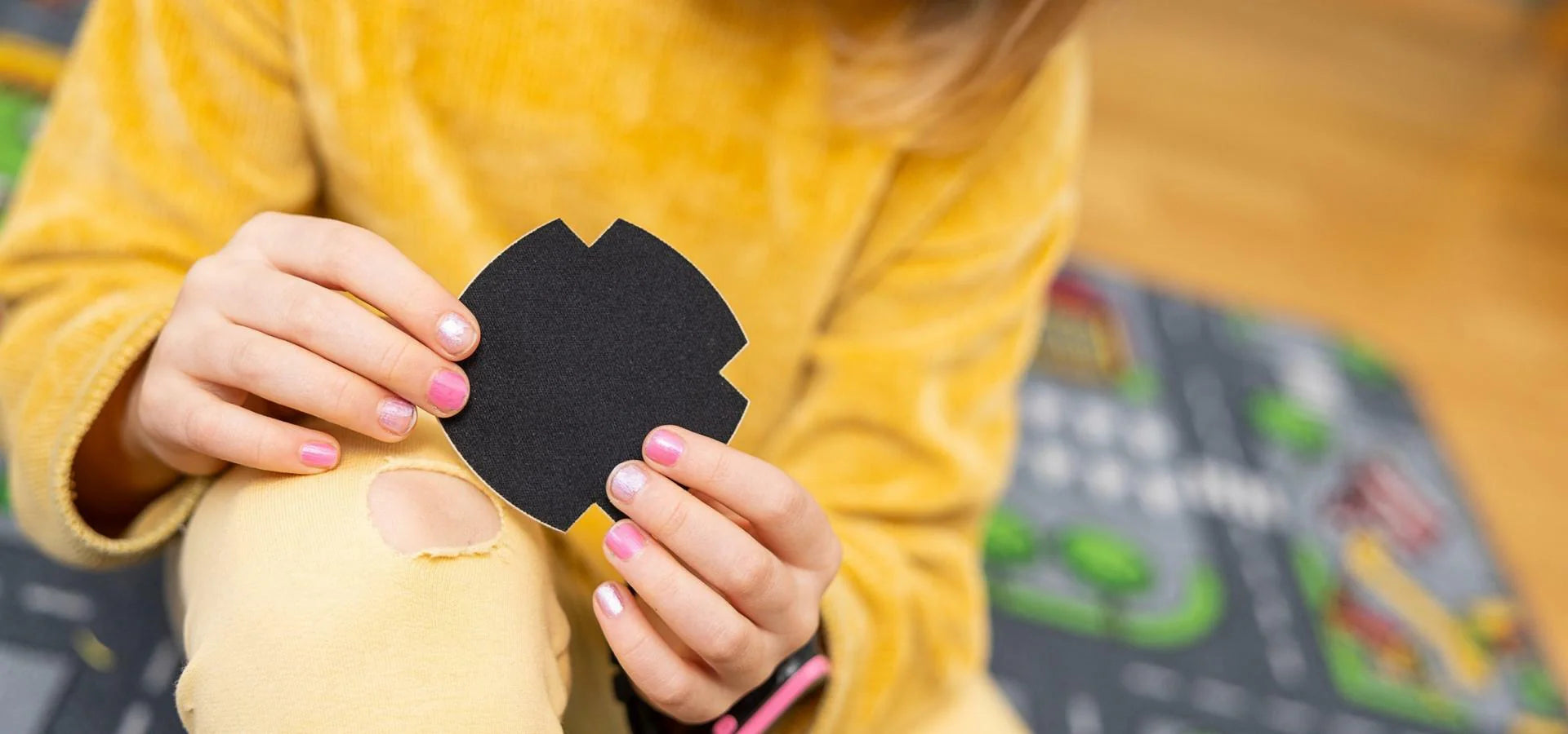 A child in a yellow outfit examines a FabPatch rub-on textile repair patch while looking at a tear in their yellow pants