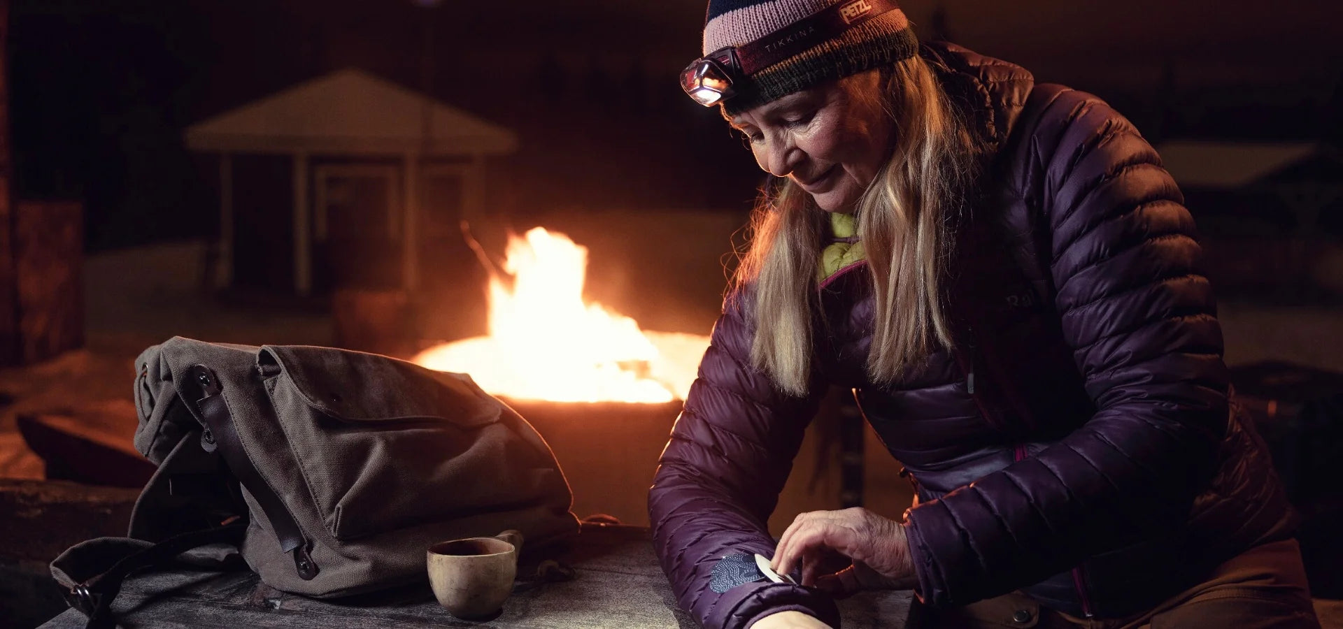 A woman wearing a beanie and headlamp applies a FabPatch rub-on textile repair patch to her puffer jacket sleeve by a campfire at night