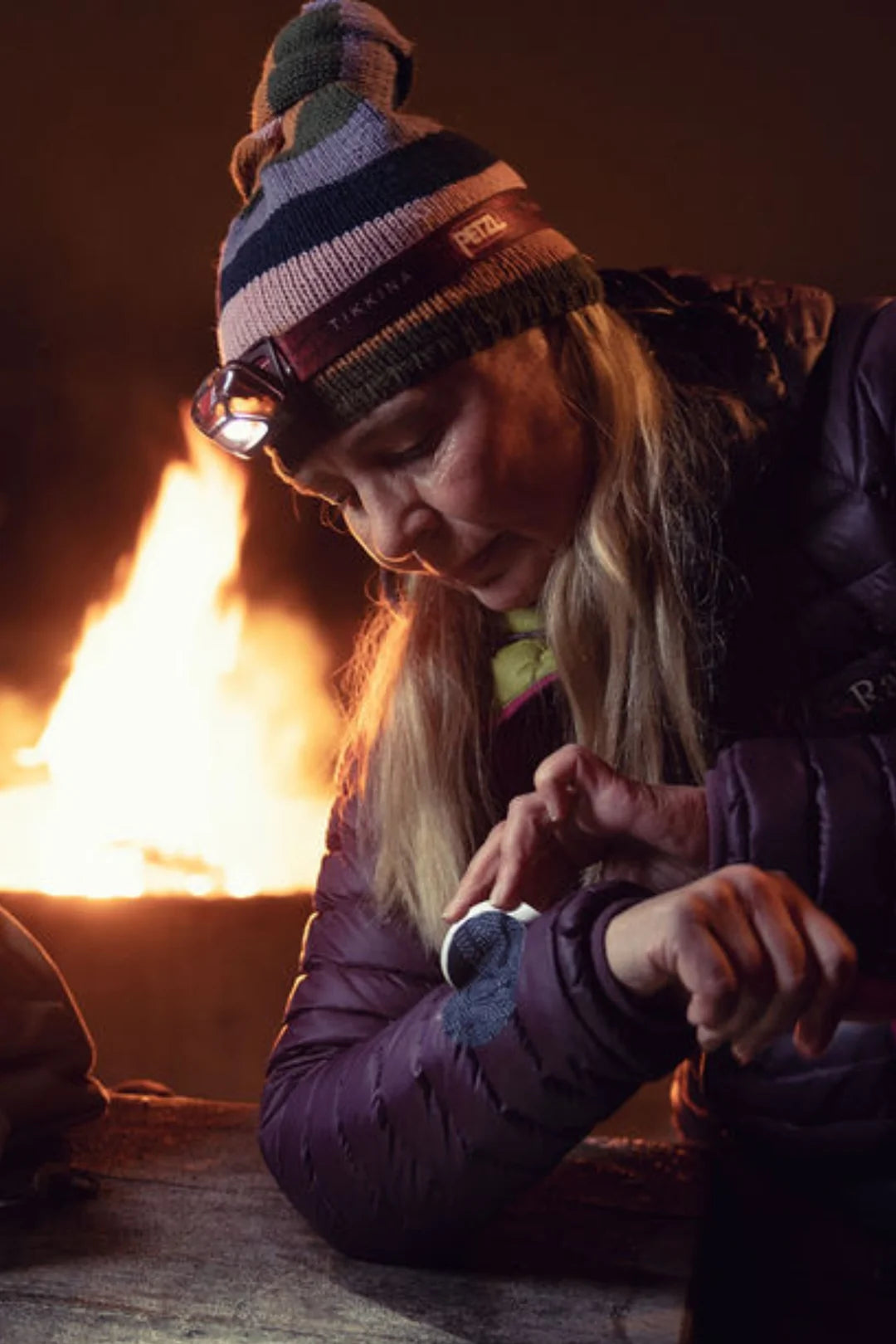 A woman wearing a beanie and headlamp applies a FabPatch rub-on textile repair patch to her puffer jacket sleeve by a campfire at night